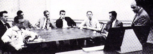 Black-and-white photo from the classic 1951 Asch social influence experiment showing one real participant seated at a table with six research confederates