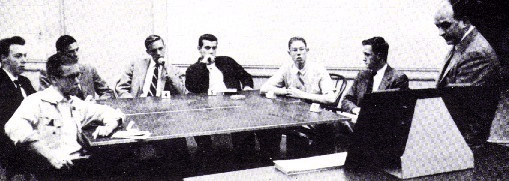 Black-and-white photo from the classic 1951 Asch social influence experiment showing one real participant seated at a table with six research confederates