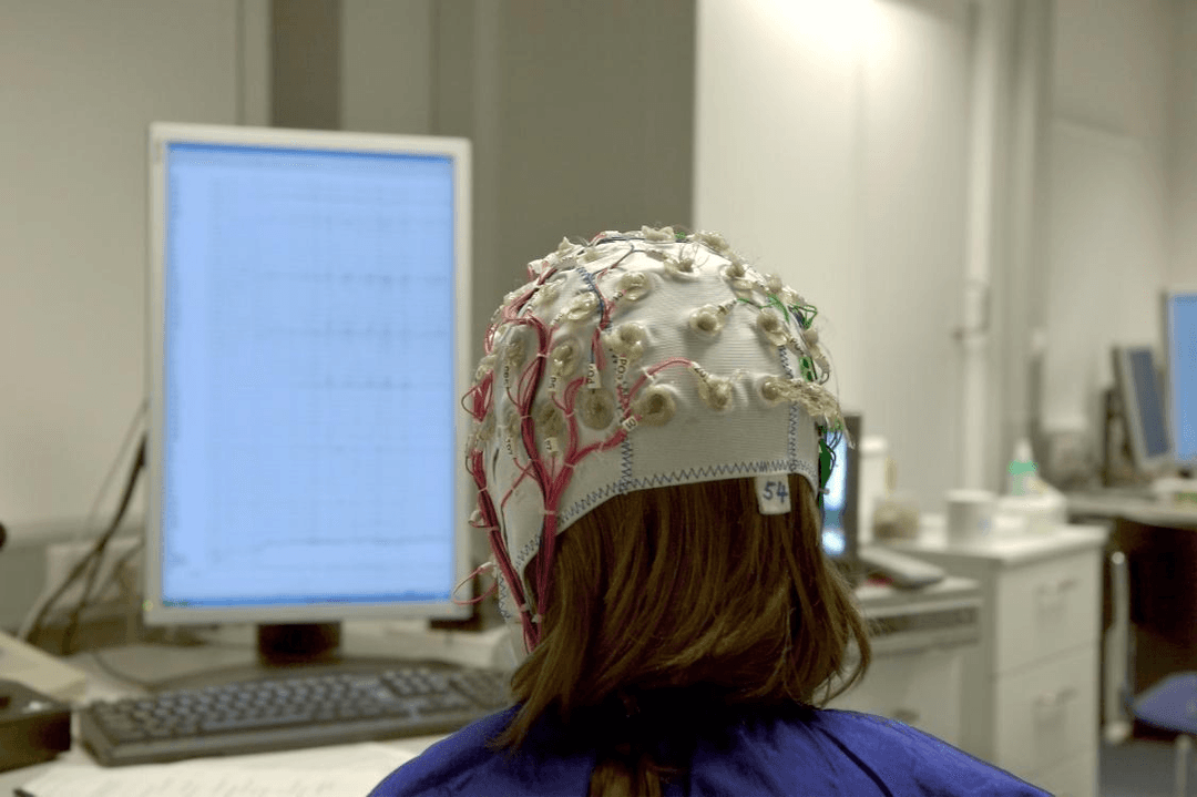 Person wearing an EEG cap with multiple electrodes attached to their scalp while working at a computer, demonstrating how electroencephalography measures brain electrical activity in behavioral research
